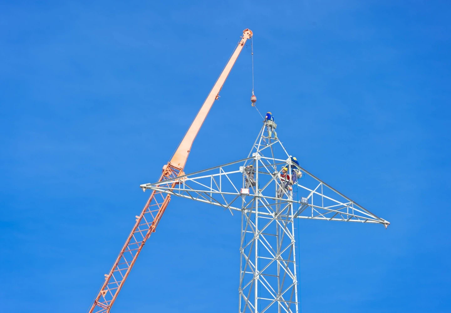 Arbeiter in Sicherheitsausrüstung montieren oder warten einen Hochspannungs-Gittermast aus Stahl mithilfe eines Krans vor einem klaren blauen Himmel.