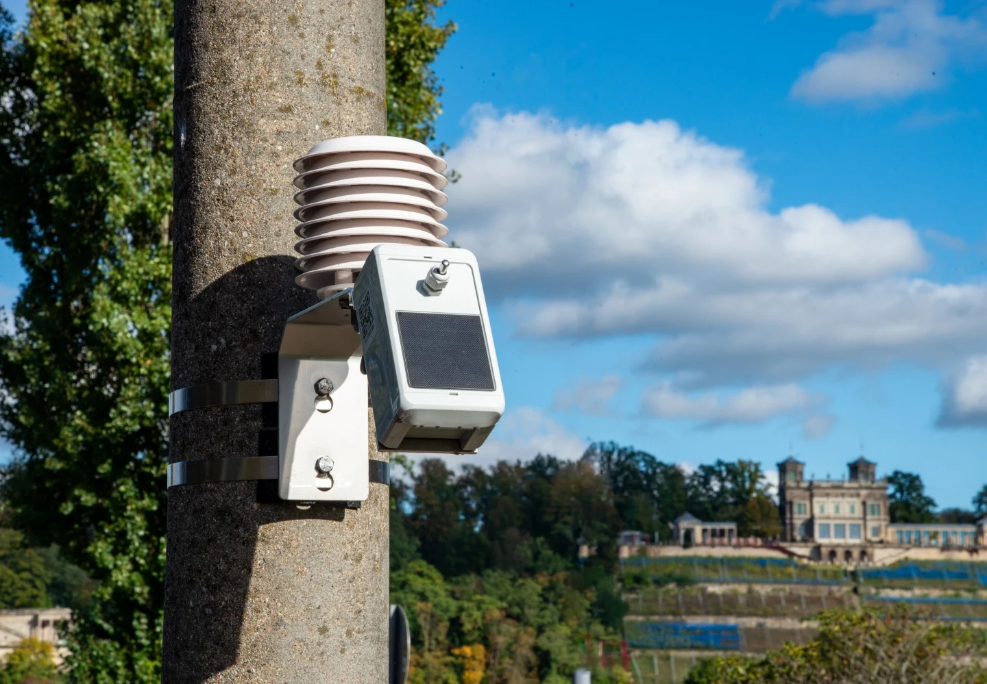 Ein Klimasensor ist an einem Laternenmast befestigt. Im Hintergrund sieht man einen Elbhang mit einem Dresdner Schloss.